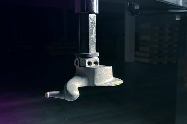 Pieza metálica montada en un vástago vertical, con forma de boquilla curvada y tres orificios visibles, iluminada lateralmente sobre fondo oscuro con palés desenfocados al fondo