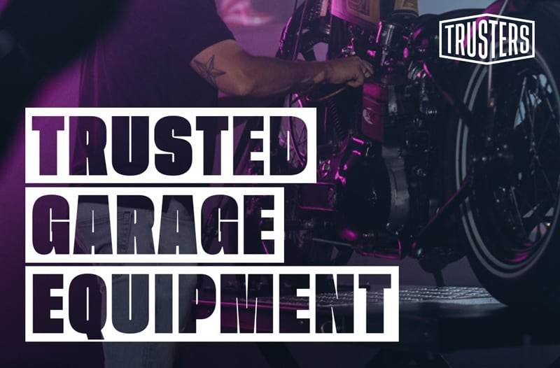 Mechaniker arbeitet an einem Motorrad auf einer Hebebühne bei lila Beleuchtung; überlagert von großen weißen Worten TRUSTED GARAGE EQUIPMENT und kleinem TRUSTERS-Logo oben rechts