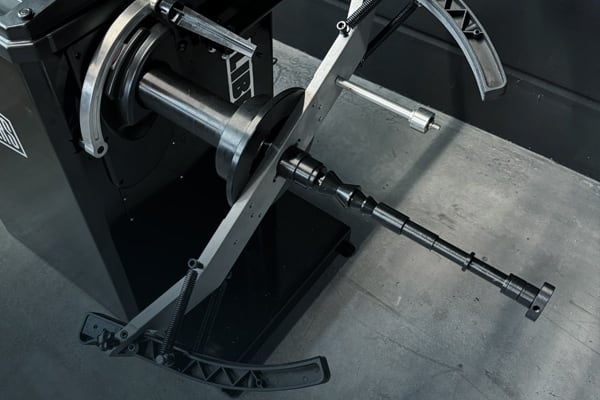 Maschinenspindel mit eingespanntem, mehrstufigem Metallwellenadapter auf einem Balancier-/Zentriergerät, umgeben von verstellbaren gebogenen Haltearmen und Zugfedern auf grauem Werkstattboden