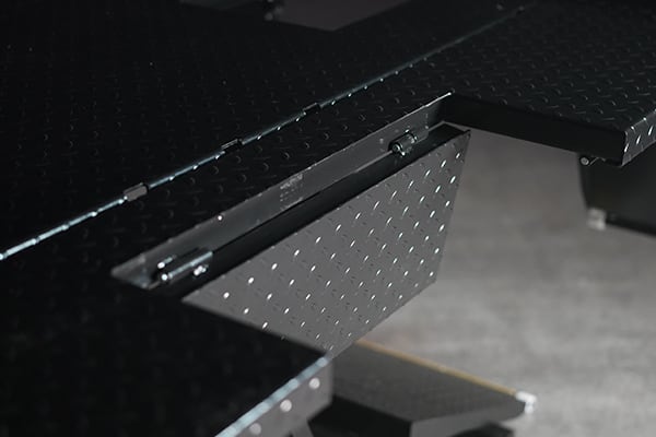 Close-up of a black diamond-plate metal platform with a partially extended hinged flap/ramp showing latch and hinge hardware, shallow depth of field and blurred concrete floor in the background
