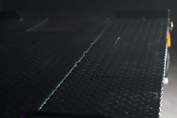Close-up of a dark metal diamond plate platform angled diagonally, showing a central seam, raised tread pattern and subtle highlights along the right edge against a dim, out-of-focus background.