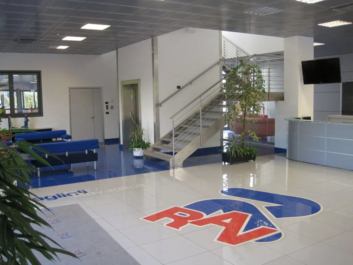Moderne helle Empfangshalle mit glänzendem Fliesenboden und großem blau-rotem RAV-Logo, Empfangstresen rechts, Treppe mit Edelstahlgeländer in der Mitte, blaue Sitzbänke links und mehrere Zimmerpflanzen