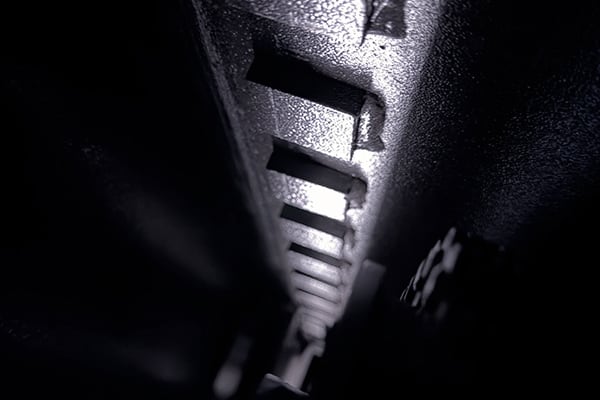 Primer plano de un raíl metálico con dientes rectangulares en perspectiva longitudinal, iluminado lateralmente que crea altos contrastes y sombras profundas, con textura rugosa y tonos gris y negro