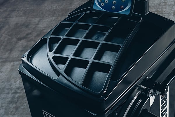 Nahaufnahme eines schwarzen Werkstattgeräts mit Ablage aus quadratischen Vertiefungen, blau beleuchtetem Bedienfeld oben und metallischem Hebel an der rechten Seite