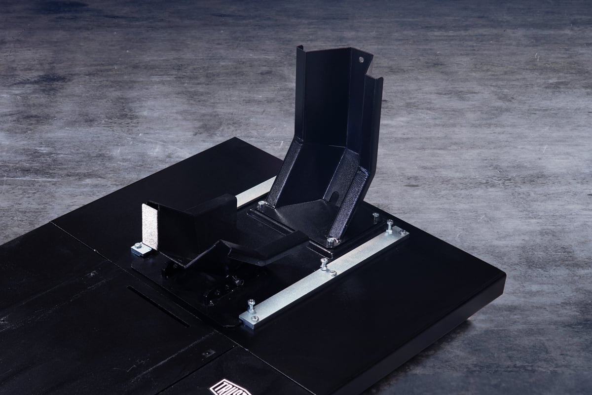 Black metal mounting bracket with angled V-shaped cradle and adjustable latch, secured to a rectangular black base with exposed bolts, photographed on a textured gray concrete floor