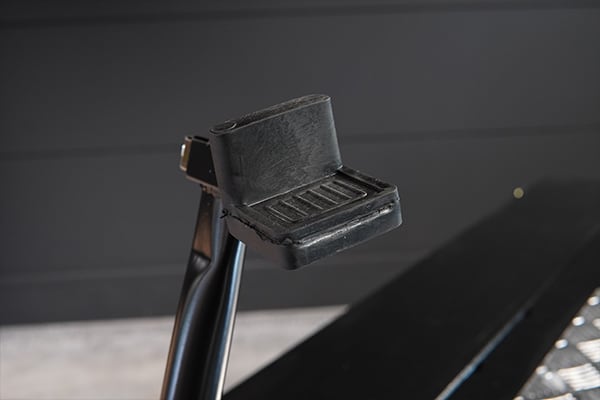 Close-up of a black plastic foot peg with a textured rectangular platform and raised heel guard, mounted on a vertical metal post against a dark gray paneled background