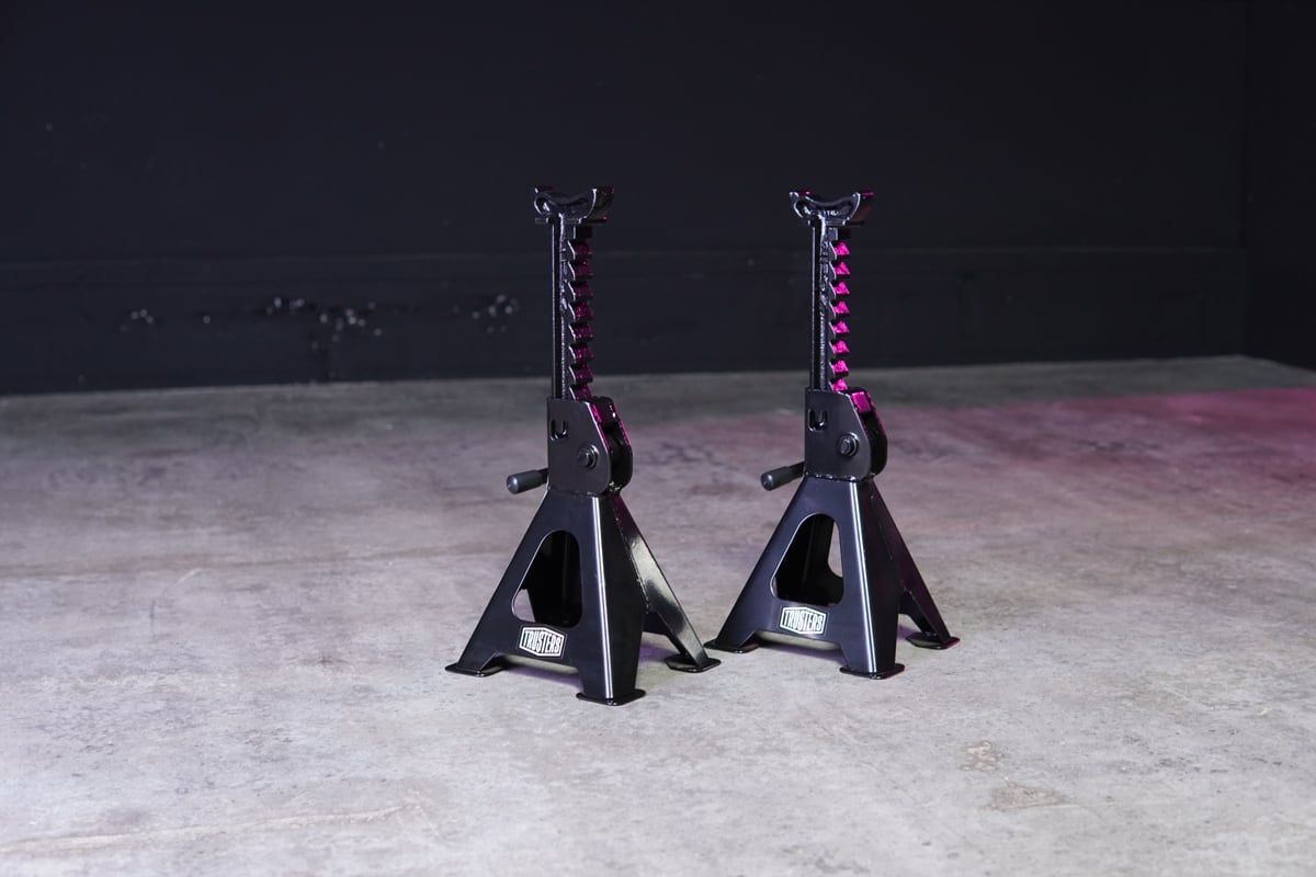 Zwei schwarze Metall-Unterstellböcke mit Zahnstangenverstellung auf Betonboden vor dunkler Wand, leichte rosa Lichtreflexe