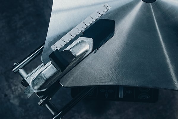 Nahaufnahme einer gebürsteten Metallplatte mit Maßstabslineal, verstellbarer Anschlagbacke und Schraubzwinge an einem Sägetisch