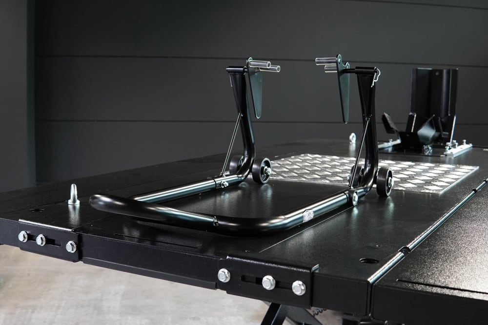 Caballete trasero para motocicleta de tubo de acero negro sobre una plataforma de trabajo negra en taller, con ruedas pequeñas, soportes en U y un tope de rueda visible al fondo