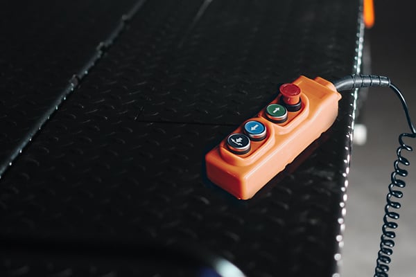 Orange industrial pendant control with three colored round buttons and a red emergency-stop button, attached coiled cable, resting on black diamond-plate metal platform.