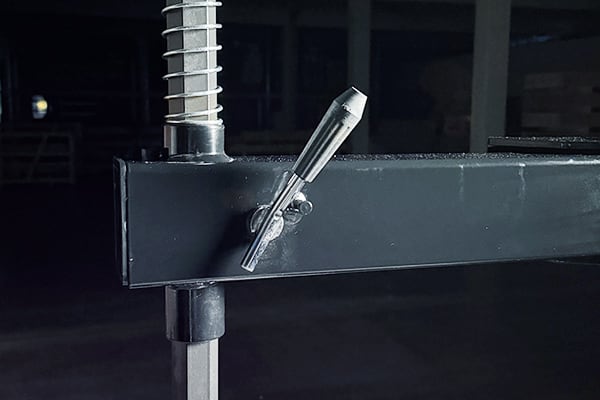 Abrazadera metálica ajustable fijada a una viga horizontal negra con perno roscado vertical, primer plano en interior oscuro de almacén