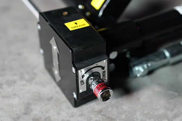 Nahaufnahme eines schwarzen Metallmechanismus mit gelbem GREASE-Aufkleber, Aufwärts-Pfeil und verstellbarer Sechskant-Schraube mit rotem Band, auf hellem Betonuntergrund liegend