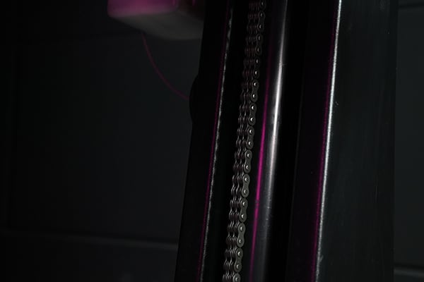 Primer plano de una cadena metálica vertical entre dos guías tubulares oscuras con reflejos púrpura y fondo en penumbra