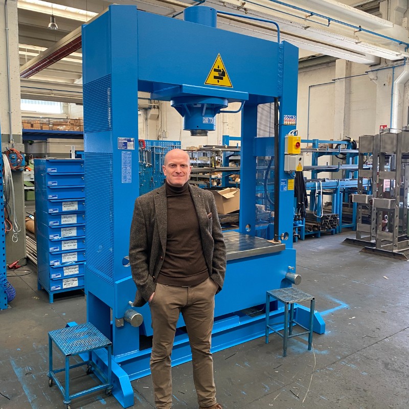 Mann vor großer blauer Hydraulikpresse in Werkhalle, trägt braunen Rollkragenpullover, graubraunes Sakko und khakifarbene Hose, Werkbänke und Regale im Hintergrund