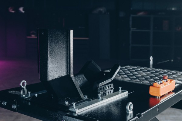 Dispositivo de sujeción metálico negro montado sobre una placa con rieles y tornillos, con anillas de enganche y un pequeño controlador naranja a la derecha, sobre una mesa de taller industrial con fondo desenfocado