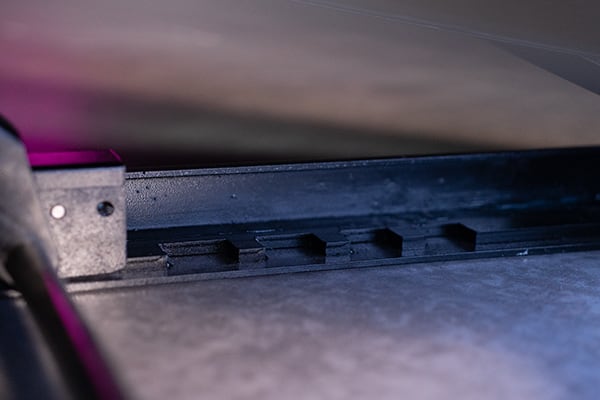 Close-up of a black metal sliding rail with rectangular notches on a gray metal surface, illuminated by soft purple and blue light