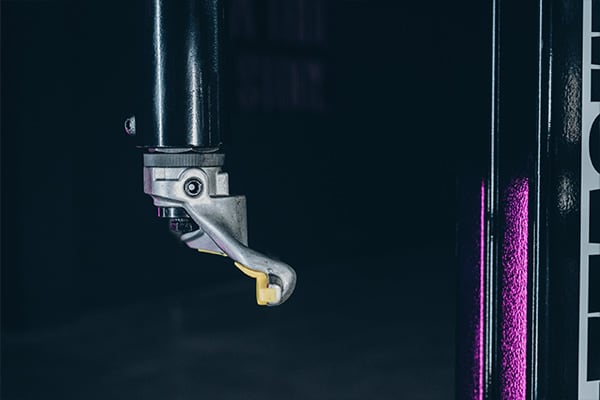 Nahaufnahme eines silbernen Metallhakens mit gelbem Kunststoffschutz am Ende, befestigt an einer schwarzen Stange; dunkler Hintergrund und rechts eine Säule mit violettem Lichtschein