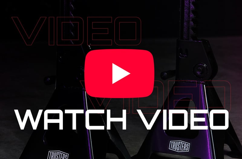 Centered red YouTube play icon above bold white text WATCH VIDEO over a dark industrial background featuring two black metal jack stands with small TRUSTERS logos and purple side lighting