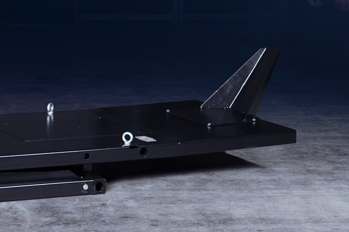 Close-up of a black metal platform with a triangular fin and two eye bolts, mounted on a scissor-like base, sitting on a concrete floor against a dark background