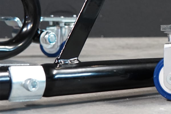 Close-up of a black tubular metal frame with welded support and silver clamp bolt, showing blue-and-gray caster wheels on a smooth concrete floor