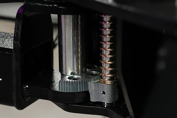 Nahaufnahme mechanischer Bauteile: glänzende Führungsstange, spiralförmige Gewindespindel, graues Rändelrad und schwarzer Metallhalter mit Befestigungslöchern und Schrauben