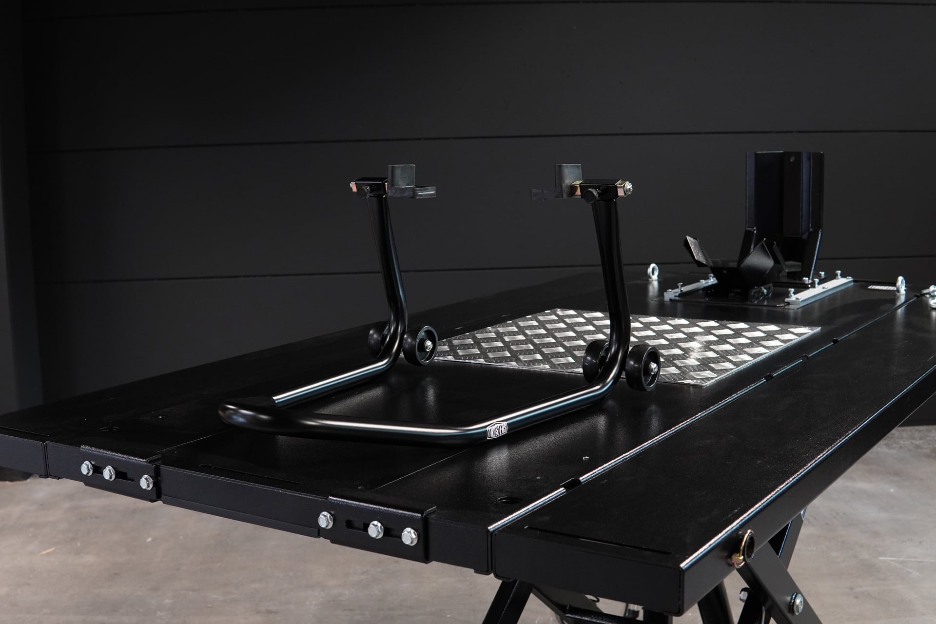 Black motorcycle rear paddock stand with twin small wheels on a black lift table, diamond plate mat and a mounted wheel chock assembly, dark paneled background