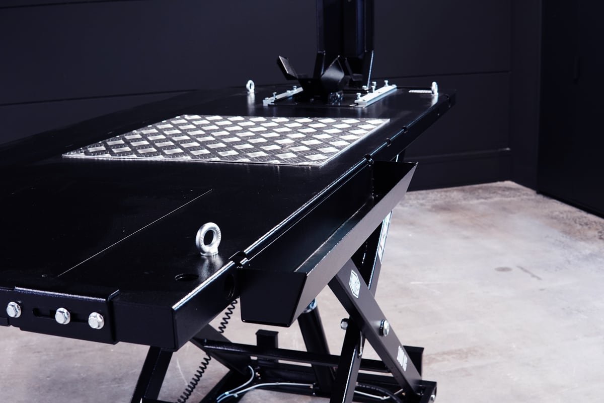 Mesa elevadora hidráulica negra tipo tijera con plataforma metálica antideslizante en patrón damero, ojales y tornillos visibles, sobre suelo de taller