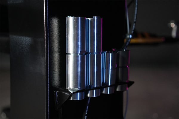 Close-up of several polished cylindrical metal tubes mounted vertically on a black support bracket, reflecting blue and purple stage lighting