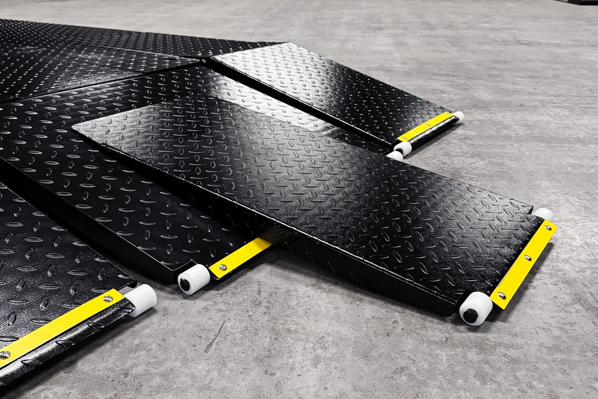 Black diamond-plate metal ramps with yellow safety edge strips and white roller wheels arranged on a grey concrete floor