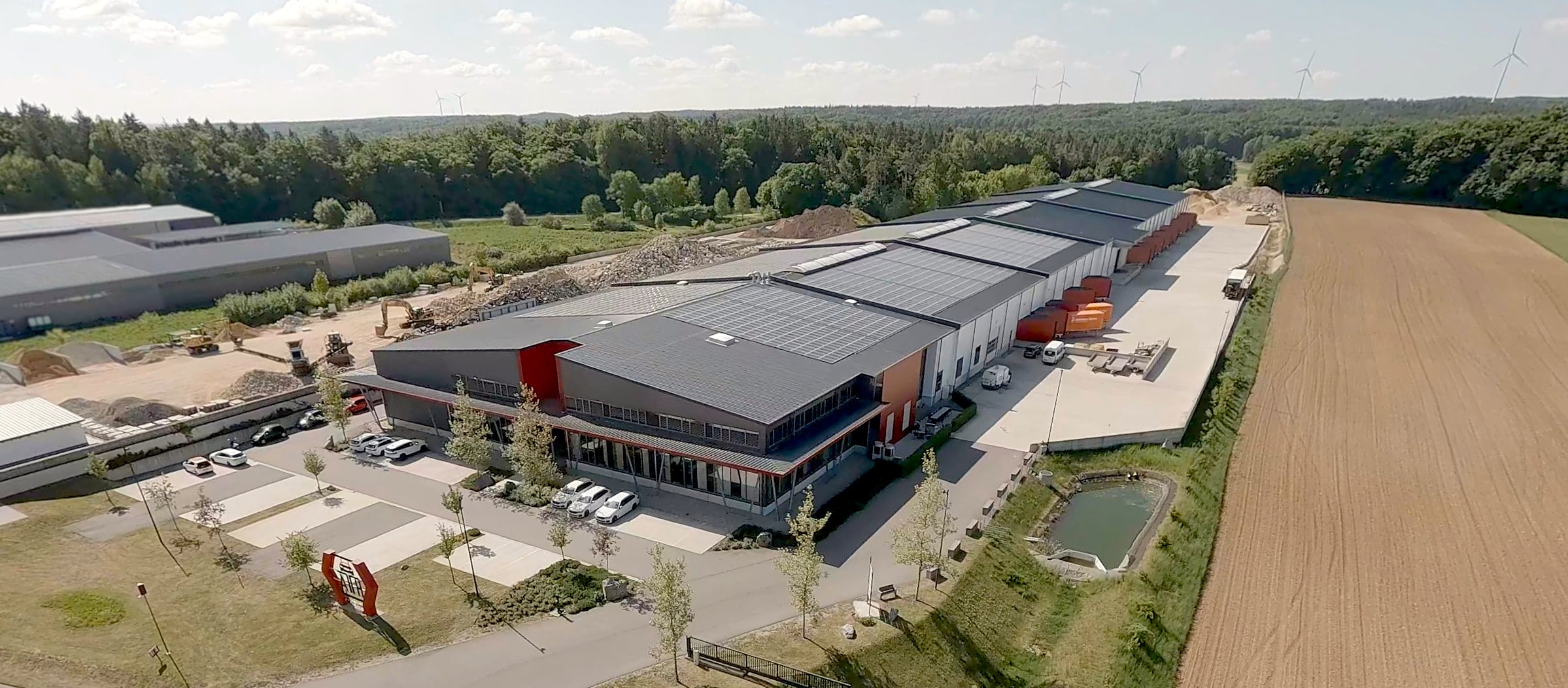 Vogelperspektive eines großen Industrie- und Lagergebäudes mit Solarpaneelen auf dem Dach, Laderampen mit orangefarbenen Anhängern, Parkplatz mit Autos, Baufahrzeugen, daneben Feld, Wald, Windräder am Horizont und kleiner Teich