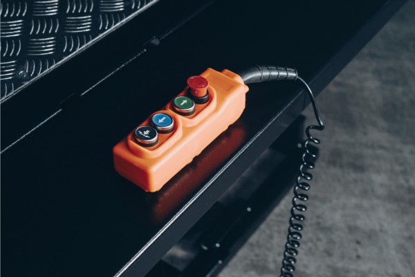 Colgante de control naranja sobre una plataforma metálica negra: mando con botón rojo de emergencia, tres botones circulares con flechas de colores y cable en espiral