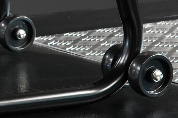 Close-up of a glossy black tubular metal frame with two small black wheels resting on a dark floor next to a textured aluminum diamond-plate surface