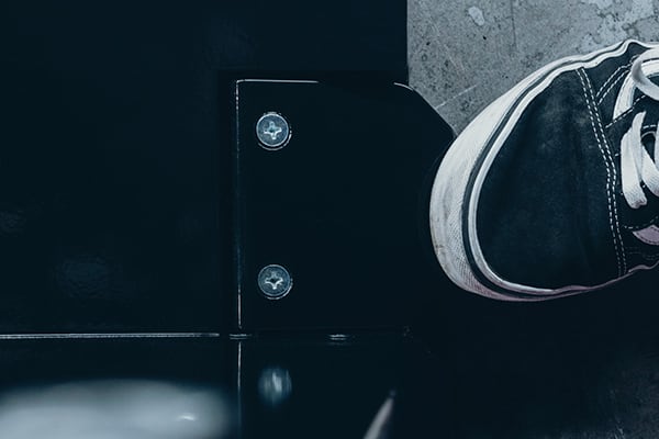 Nahaufnahme eines schwarzen Sneakers mit weißen Schnürsenkeln neben einer glänzenden schwarzen Metallkante mit zwei Schrauben auf hellem Betonboden