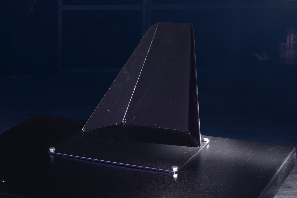 Black metal fin-shaped bracket mounted on a square base plate with visible hex bolts, photographed against a dark studio background
