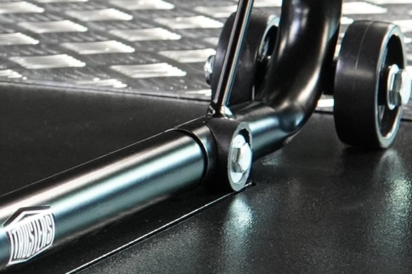 Primer plano de soporte metálico negro con tubo, tornillo de cabeza hexagonal, rueda pequeña de goma y placa de acero acanalada de fondo