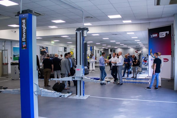 Helle Werkstatthalle mit mehreren Fahrzeughebebühnen, blauen Säulen mit Logo und Bannern; zahlreiche Personen in kleinen Gruppen unterhalten sich und gehen durch den offenen Messestand- und Servicebereich