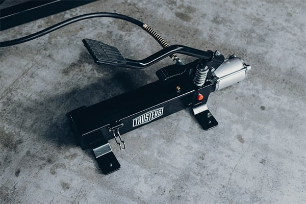 Black foot-operated hydraulic pump with metal pedal, attached hose and silver cylinder actuator, visible spring and red release button, TRUSTERS logo on the side, placed on a stained concrete floor