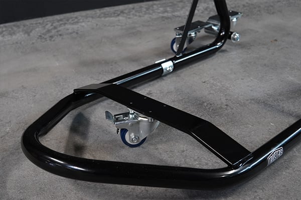 Close-up of a black metal motorcycle paddock stand showing its tubular frame, metal support bracket and small blue swivel caster wheels resting on a concrete floor
