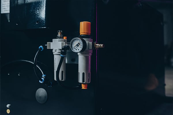 Druckluftfilter und -regler mit Manometer, zwei grauen Filtergehäusen, orangefarbenen Einstellknöpfen und angeschlossenen Schläuchen an einem schwarzen Maschinengehäuse