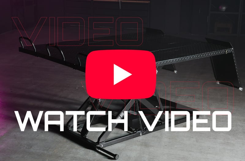 Black hydraulic scissor lift table on a concrete warehouse floor with a red YouTube play button overlay and large white text reading WATCH VIDEO across the foreground