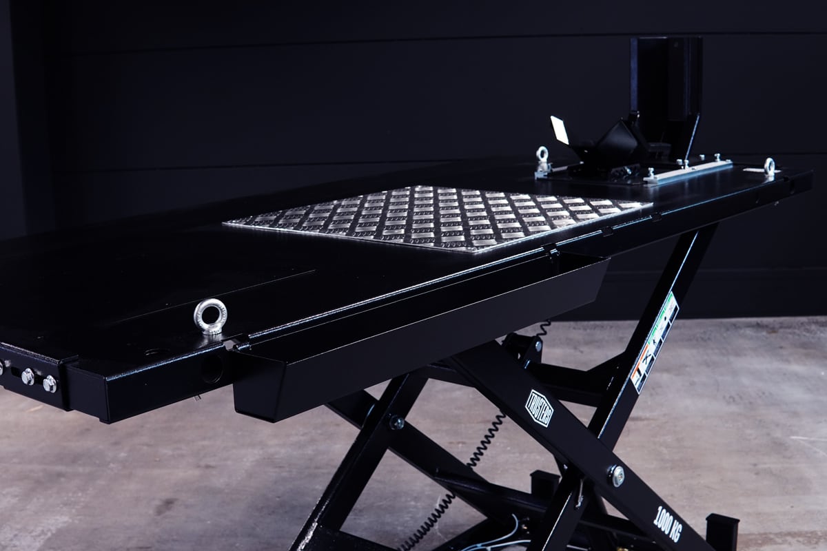 Mesa elevadora negra para motos tipo tijera con plataforma de placa de diamante, anillas de sujeción, mecanismo hidráulico visible y etiqueta 1000 KG, sobre suelo de hormigón y fondo oscuro