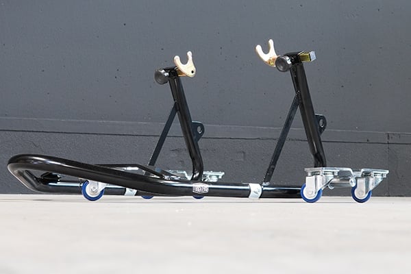 Two black motorcycle paddock stands with white fork hooks and blue caster wheels resting on a concrete floor against a gray wall
