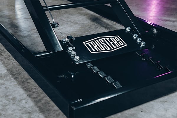 Close-up of a black metal hinge plate and bolted frame on a concrete floor, featuring a rectangular label reading THUSTERS and subtle purple light reflections on the glossy surface