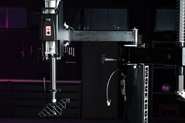 Prensa de taladro industrial negro con brazo horizontal y accesorio de sujeción metálico en primer plano, banco de trabajo oscuro y estanterías al fondo con iluminación morada tenue