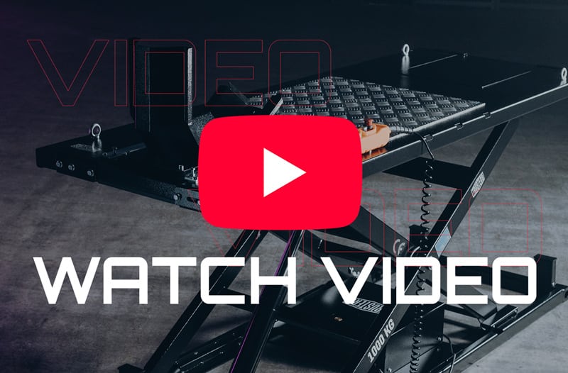 Black scissor lift platform in a workshop with diamond-plate surface, coiled remote cable and orange handheld controller; overlaid large red YouTube play button graphic and bold white text reading WATCH VIDEO
