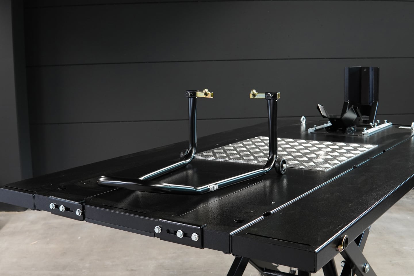 Black tubular motorcycle rear paddock stand with gold hooks and small wheels resting on a black hydraulic lift table beside a diamond-plate foot panel and front wheel chock in a dim workshop