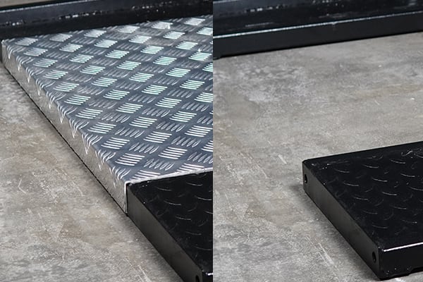 Dos piezas metálicas con relieve antideslizante sobre un suelo de hormigón: a la izquierda una rampa inclinada de chapa de aluminio plateada con patrón de rombos, a la derecha un bloque recto negro con el mismo dibujo.