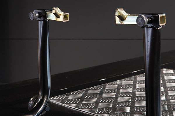 Two black tubular metal supports with brass-colored square clamp fittings mounted above a silver diamond-plate metal surface on a dark background