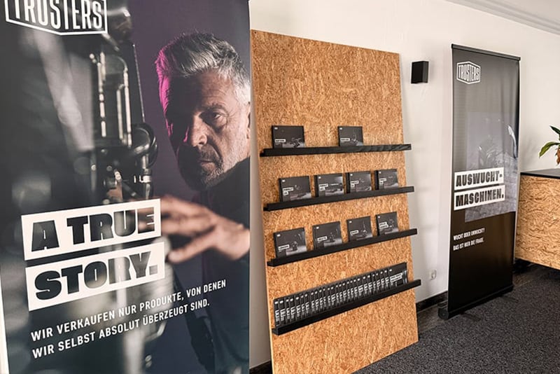 Showroom-Ecke mit zwei Rollup-Bannern: links Portrait eines Mannes und Schriftzug A TRUE STORY, rechts Banner mit Schriftzug AUSWUCHT-MASCHINEN; daneben eine Holztafel mit Regalbrettern und Reihen kleiner Produktverpackungen