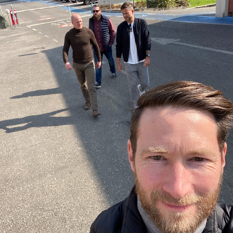 Selfie eines lächelnden Mannes im Vordergrund; drei weitere Männer gehen hinter ihm über einen asphaltierten Parkplatz bei Sonnenschein, alle in lässiger Alltagskleidung